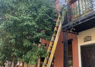 UMAR Painting Co. painter on an extension ladder painting the upper window trim and cornice of a terracotta Philadelphia, PA rowhome with a black wrought iron balcony