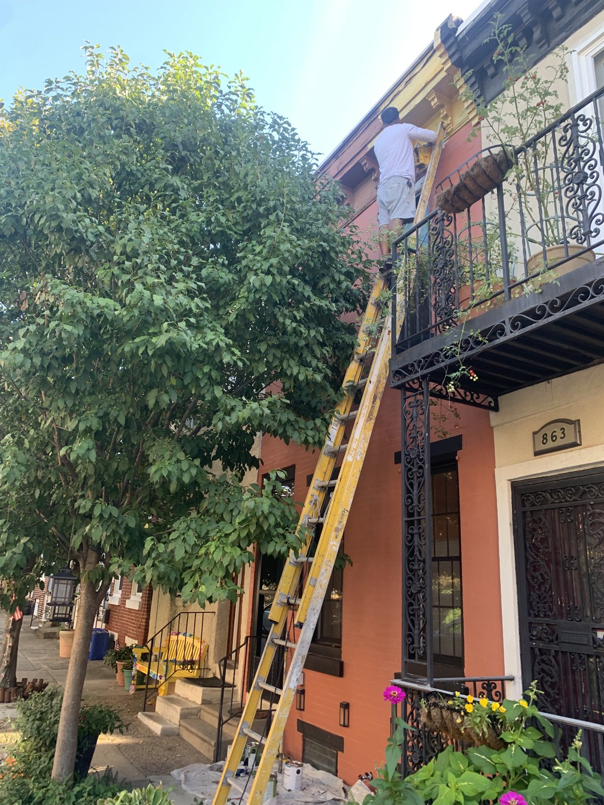 UMAR Painter on Ladder Painting Rowhome Exterior Trim, Philadelphia, PA | Affordable Philly Painters UMAR Painting Co. painter on an extension ladder painting the upper window trim and cornice of a terracotta Philadelphia, PA rowhome with a black wrought iron balcony